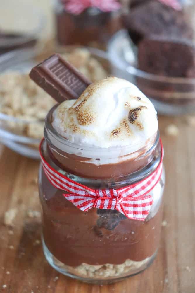 A jar of chocolate mousse topped with toasted marshmallow and a piece of chocolate, adorned with a red and white striped ribbon. In the background, pieces of chocolate cake and crushed cookies are visible on a wooden surface.