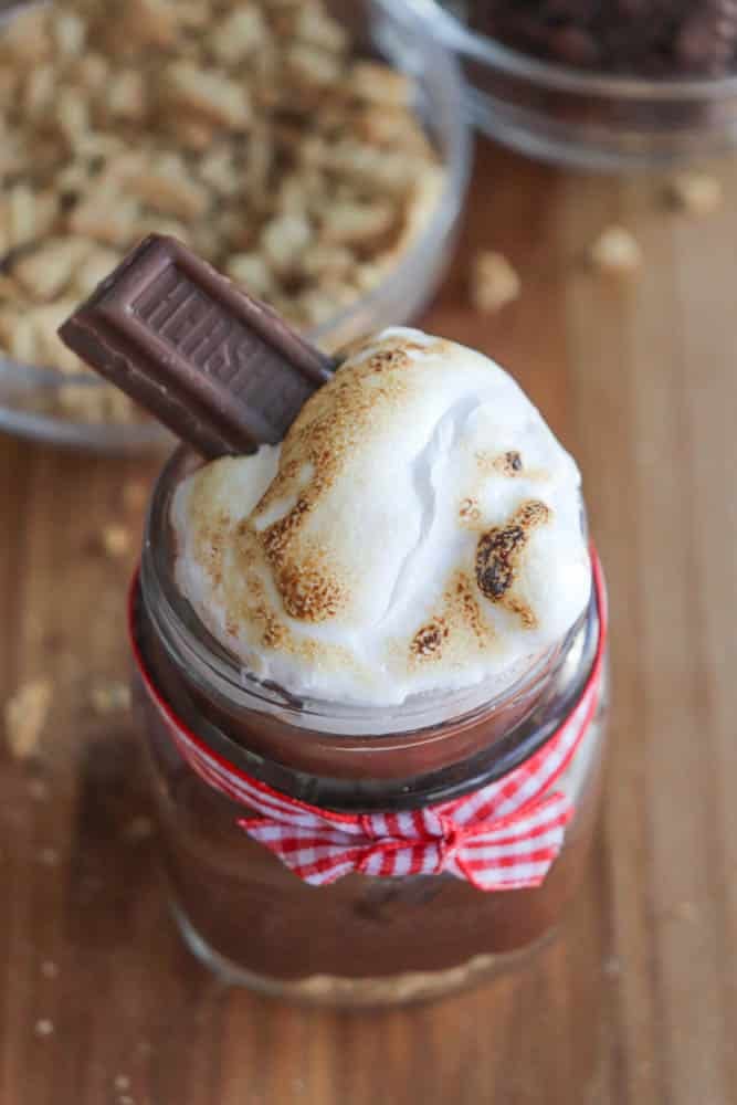 A jar dessert topped with toasted marshmallow and a piece of chocolate bar, wrapped with a red gingham ribbon. Crushed graham crackers are visible in the background on a wooden surface.