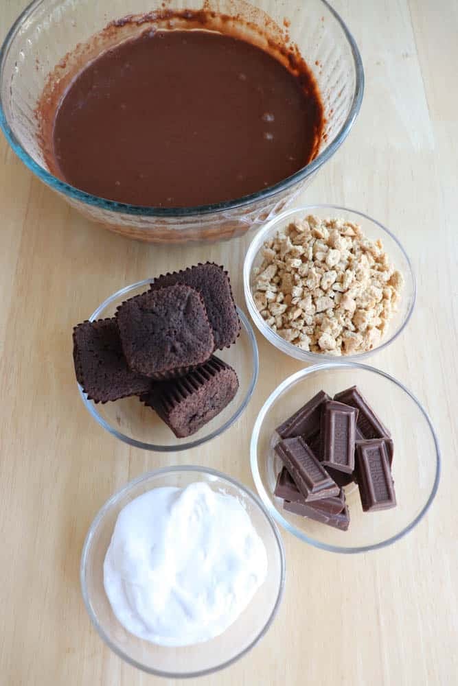 A bowl of chocolate batter is surrounded by smaller bowls containing whipped cream, chocolate bar pieces, granola, and chocolate muffins, all set on a light wood surface.