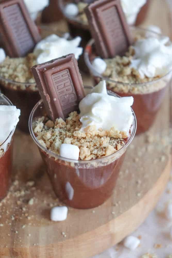 A dessert cup filled with rich chocolate pudding, topped with crumbled graham crackers, a dollop of whipped cream, and a piece of chocolate. Small marshmallows scattered around the cup on a wooden surface complete these delightful smores pudding shots.