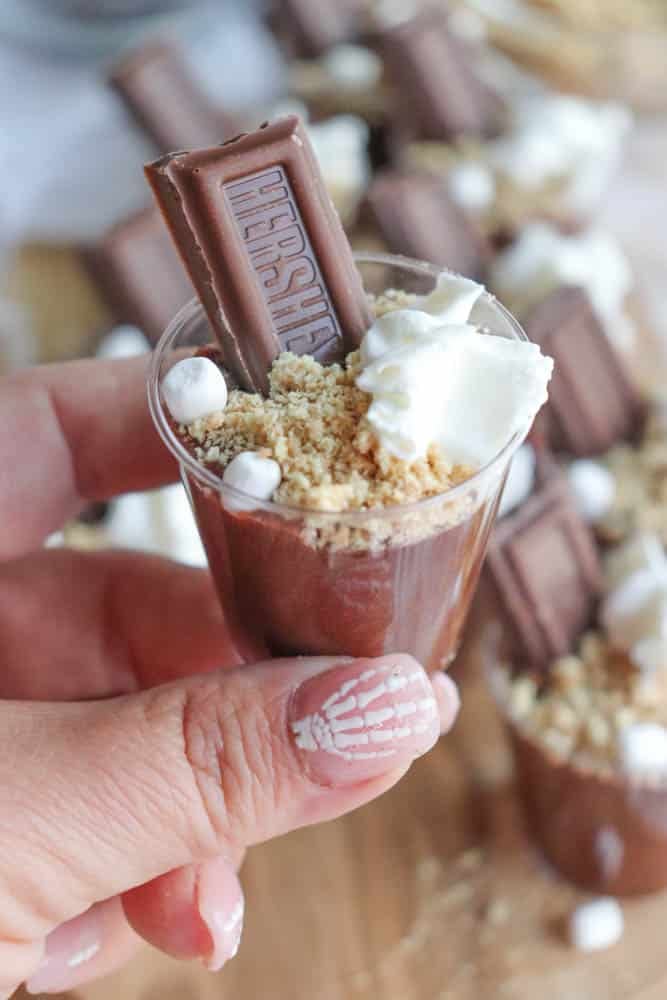 A hand holds a mini cup of smores pudding shot, filled with chocolate pudding and topped with crumbled graham crackers, whipped cream, and a piece of Hersheys chocolate. The nails show off a white and silver geometric pattern. More cups are visible in the background.