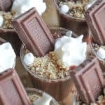 Small cups filled with chocolate mousse are topped with dollops of whipped cream, crushed graham crackers, and a piece of chocolate bar, creating an enticing presentation reminiscent of smores pudding shots.