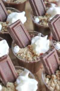 Small cups filled with chocolate mousse are topped with dollops of whipped cream, crushed graham crackers, and a piece of chocolate bar, creating an enticing presentation reminiscent of smores pudding shots.