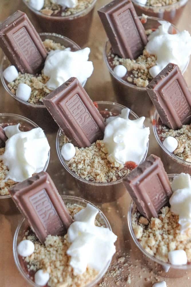 Small dessert cups filled with chocolate pudding transform into delightful smores pudding shots, topped with crushed graham crackers, a chocolate bar piece, mini marshmallows, and whipped cream, arranged in neat rows on a tray.