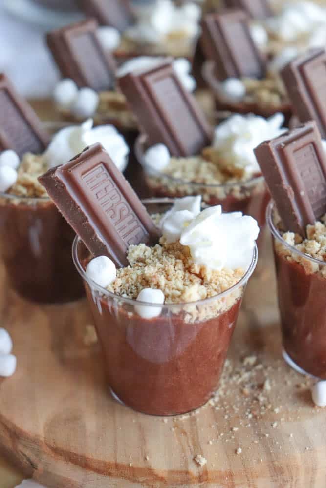 Small cups filled with chocolate pudding, transforming into delightful smores pudding shots with the addition of graham cracker crumbs, a piece of chocolate, mini marshmallows, and a dollop of whipped cream, all artfully arranged on a rustic wooden surface.