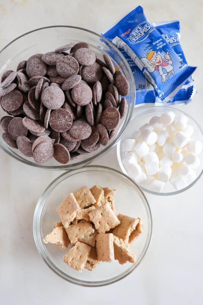 Bowls containing chocolate wafers, mini marshmallows, and graham cracker pieces are arranged on a counter. Two blue packets of marshmallow pies are in the background.