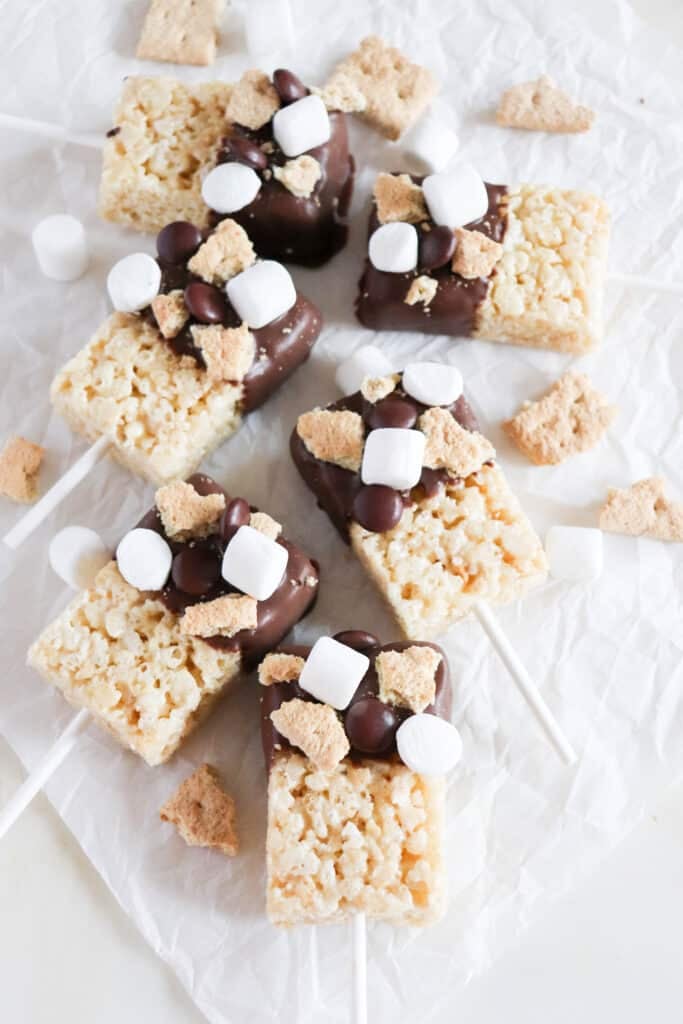 Five Rice Krispies treats on sticks, partially dipped in chocolate, topped with mini marshmallows, chocolate candies, and cookie pieces. They are arranged on crumpled parchment paper with additional toppings scattered around.