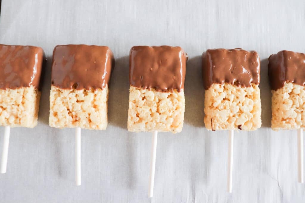 Five rectangular Rice Krispie treats on sticks, lined in a row. The top half of each treat is dipped in milk chocolate, creating a shiny coating. They are arranged on a light gray surface.