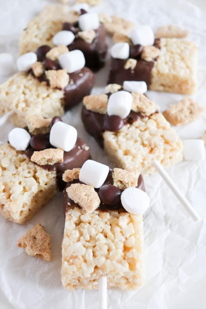 Rice Krispie treats on sticks, dipped in chocolate and topped with mini marshmallows and graham cracker pieces, placed on white parchment paper.