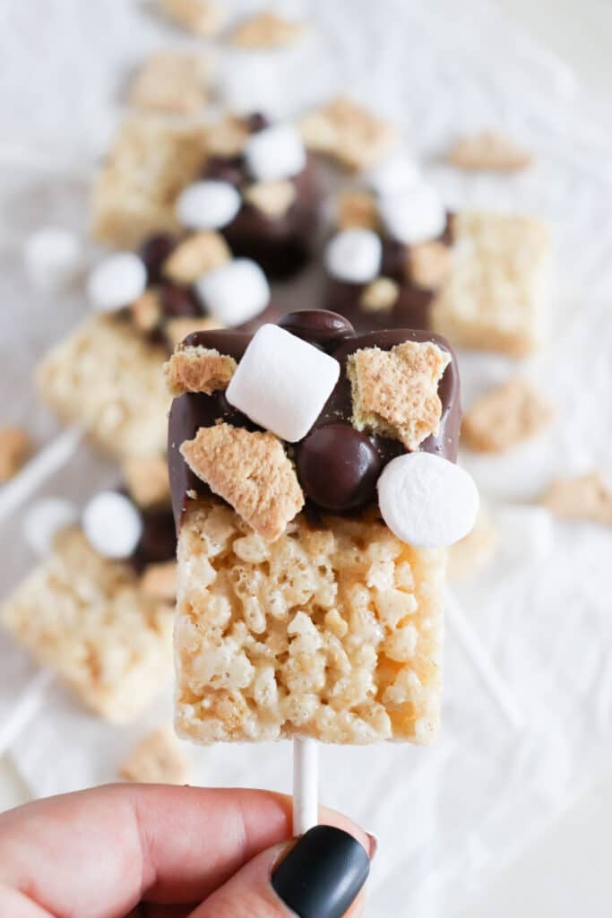 A hand with black nail polish holds a Rice Krispies treat on a stick, topped with chocolate, mini marshmallows, and graham cracker pieces. More treats are blurred in the background.