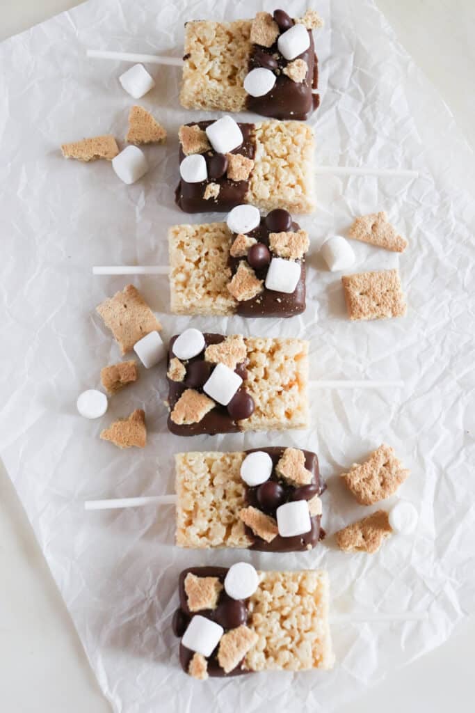 Rice Krispies treats on sticks are topped with chocolate, mini marshmallows, and graham cracker pieces, arranged in a neat row on crinkled parchment paper with some scattered toppings around.