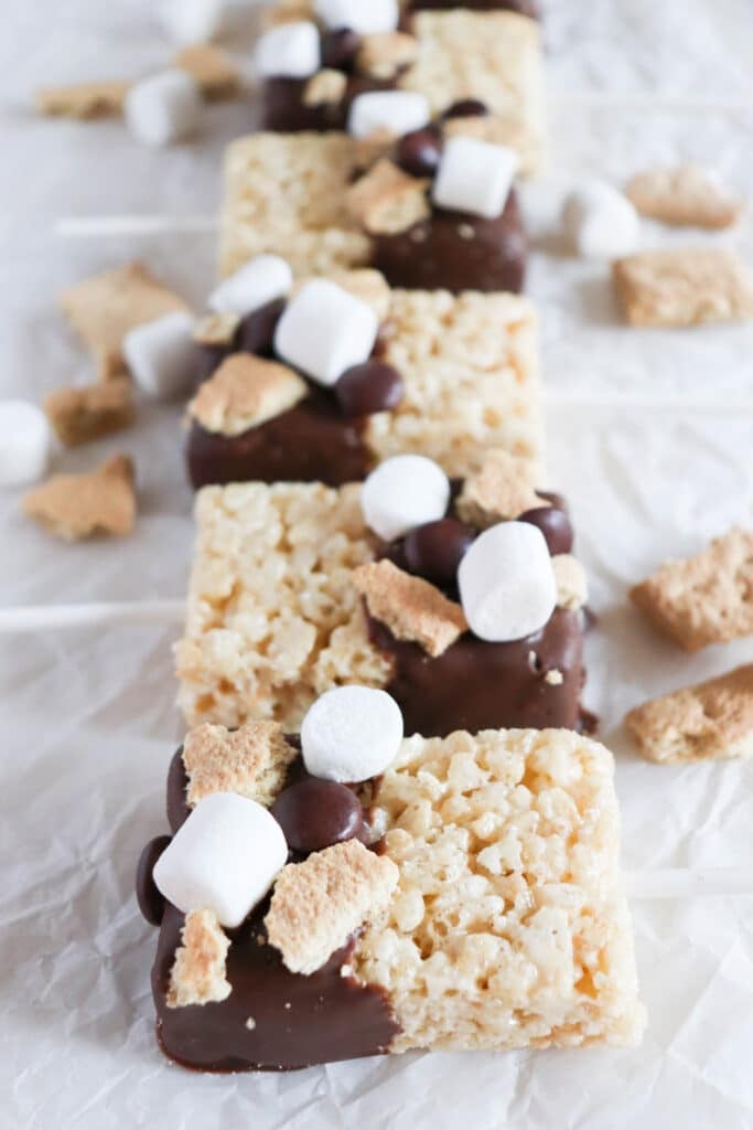 Rice Krispie treats partially dipped in chocolate, topped with mini marshmallows, chocolate chips, and graham cracker pieces, arranged in a row on crinkled parchment paper. Scattered marshmallows and graham crackers are visible around them.