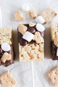 Rice Krispie treats on sticks are dipped in chocolate and topped with mini marshmallows, chocolate candies, and graham cracker pieces. They are placed on crinkled parchment paper with more treats and toppings scattered around.