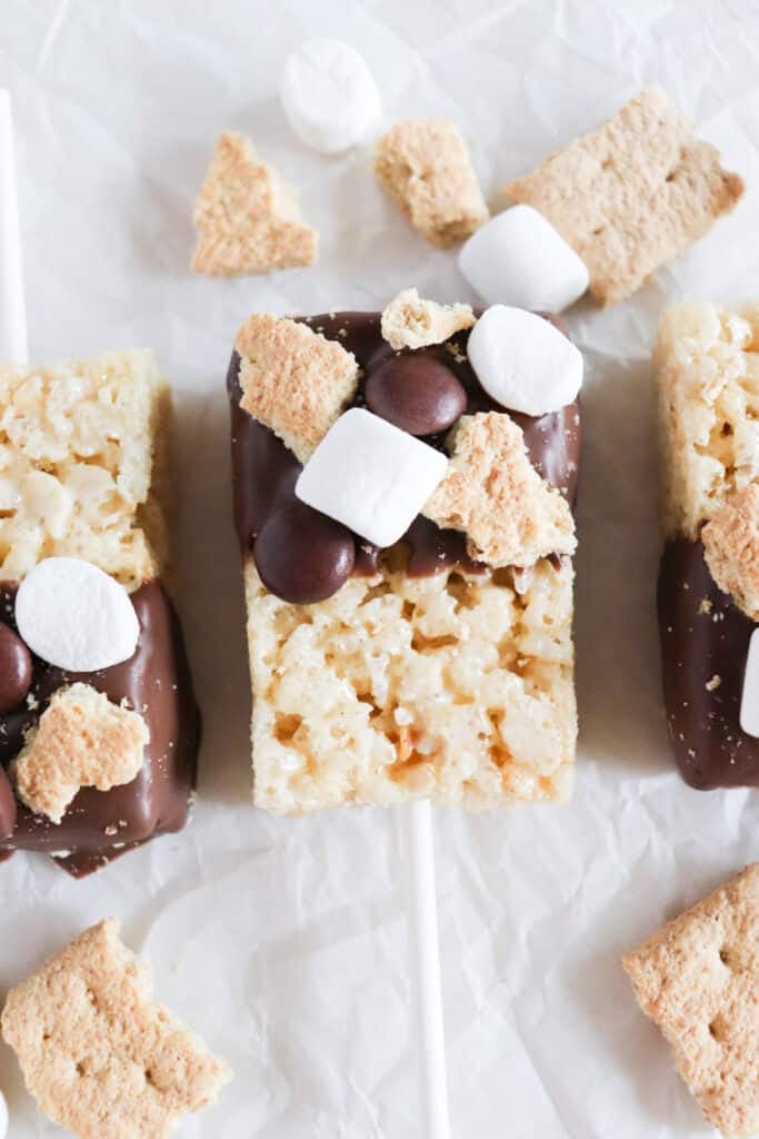 Rice Krispie treats on sticks, half-dipped in chocolate, topped with mini marshmallows, graham cracker pieces, and chocolate candies, are arranged on crumpled white parchment paper.