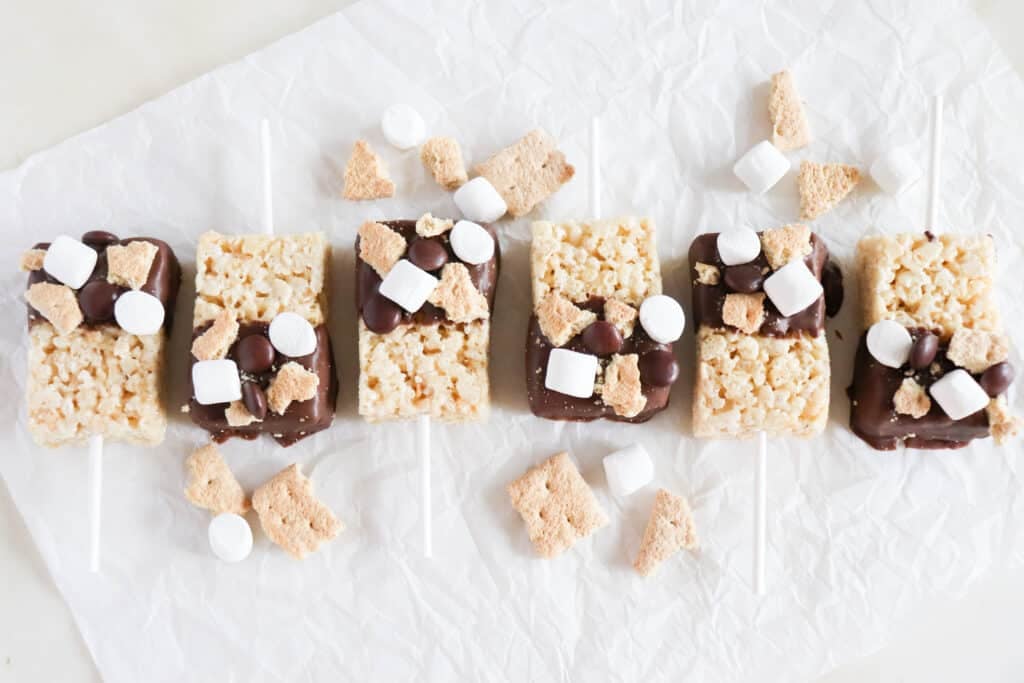 Five rice cereal treats on sticks are partially dipped in chocolate and topped with mini marshmallows, chocolate candies, and graham cracker pieces. They are arranged in a row on crinkled parchment paper.