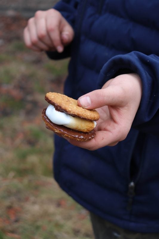 A person in a blue jacket holds what might just be the best smore, featuring a toasted marshmallow and chocolate nestled between two cookies. The grassy background is sprinkled with scattered brown leaves.