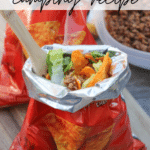 A small bag of Doritos is opened and filled with taco ingredients like lettuce, ground beef, and cheese. A wooden spoon is inside the bag, and more taco ingredients are visible in bowls in the background.