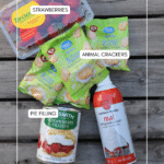 A flat lay of strawberries, a can of strawberry pie filling, several packs of animal crackers, and a can of whipped cream, all labeled, on a wooden surface.