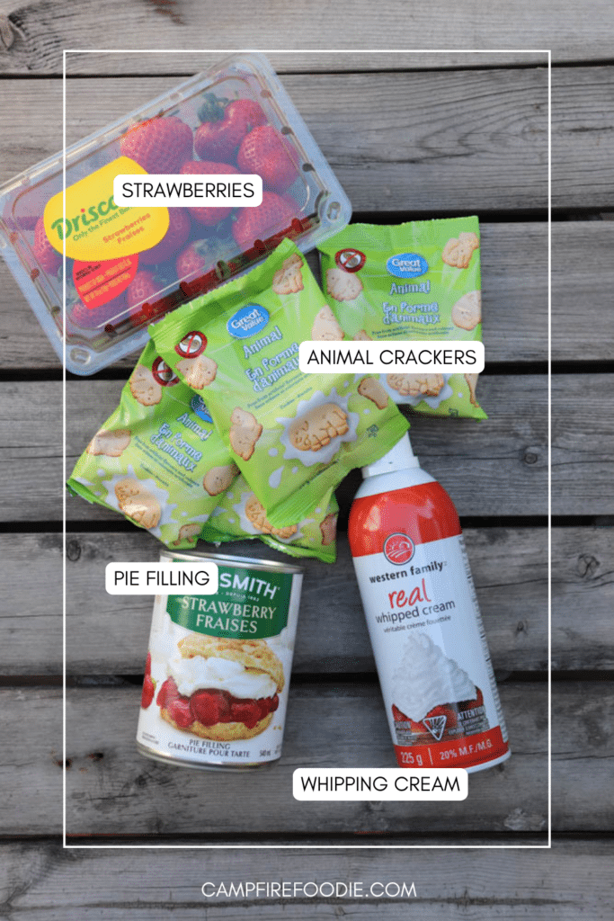 A flat lay of strawberries, a can of strawberry pie filling, several packs of animal crackers, and a can of whipped cream, all labeled, on a wooden surface.