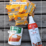 Four packs of Bear Club Grahams, a can of pumpkin pie filling, and a can of whipped cream are laid out on a wooden surface. Each item is labeled with white text.