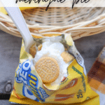 A bag of mini Golden Oreo cookies is opened and filled with lemon meringue pie ingredients, topped with whipped cream and a cookie, with a plastic spoon inside. The image also shows a picnic basket and a plaid cloth.