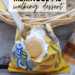 A small bag of golden Oreo cookies is opened and filled with whipped cream and cookie pieces, with a plastic spoon inside. The bag sits in a basket next to a plaid cloth. Text reads Lemon Meringue Pie walking dessert.