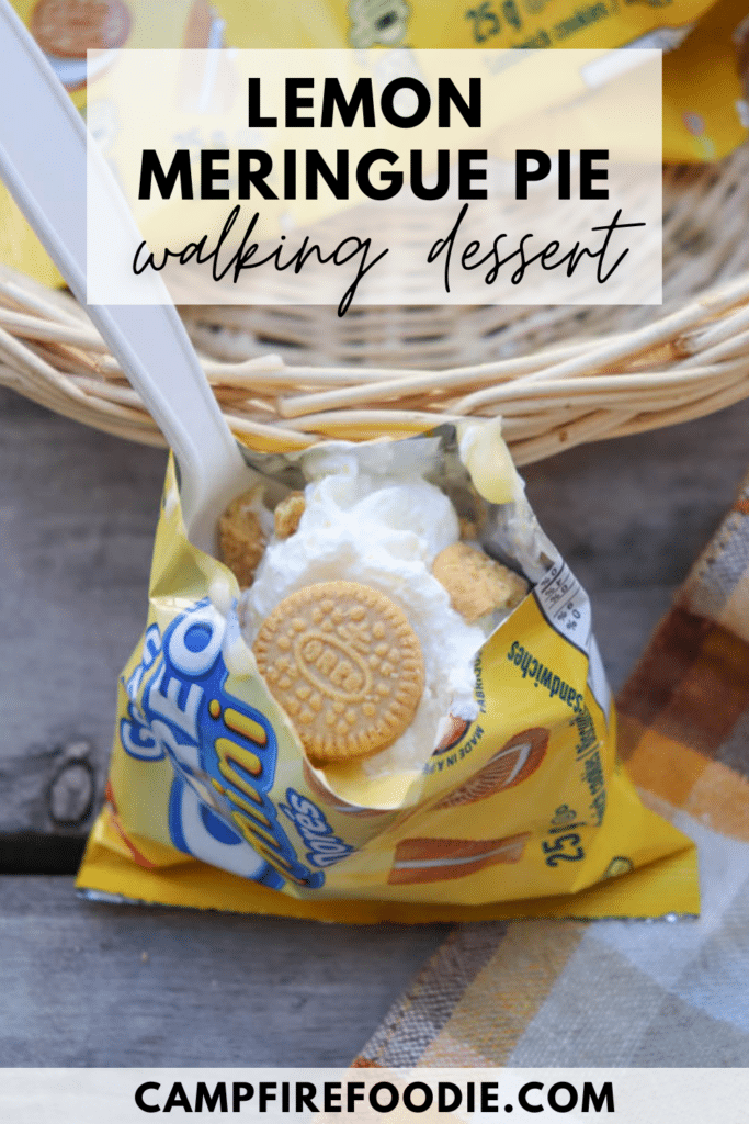 A small bag of golden Oreo cookies is opened and filled with whipped cream and cookie pieces, with a plastic spoon inside. The bag sits in a basket next to a plaid cloth. Text reads Lemon Meringue Pie walking dessert.
