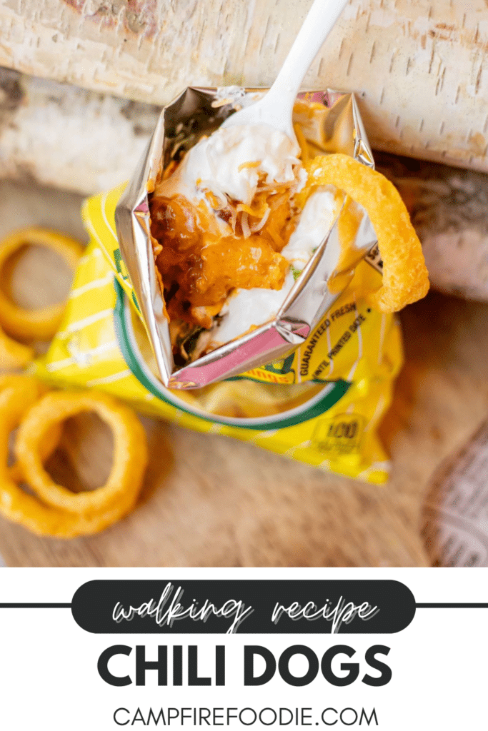 An open bag of corn chips filled with chili, cheese, and sour cream, topped with a plastic fork, sits next to crispy onion rings on a wooden surface. Text below reads: walking recipe CHILI DOGS campfirefoodie.com.