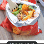 A small bag of tortilla chips is opened and filled with taco ingredients like ground beef, lettuce, and cheese. A wooden fork is inside the bag, and a label below reads “Camping Recipe: Walking Tacos, campfirefoodie.com.”.