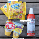Three packs of Golden Oreo Mini cookies, a can of whipped cream, and a lemon meringue pudding cup on a wooden surface, each item labeled with white text boxes.