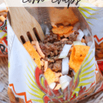 Open corn chip bag filled with ground beef, shredded cheese, diced onions, and chips. A wooden fork is inside the bag, with the text Walking Taco Corn Chips at the top and campfirefoodie.com at the bottom.