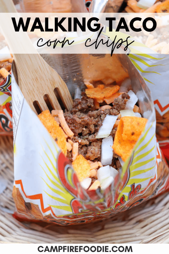 Open corn chip bag filled with ground beef, shredded cheese, diced onions, and chips. A wooden fork is inside the bag, with the text Walking Taco Corn Chips at the top and campfirefoodie.com at the bottom.
