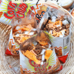 Three open bags of corn chips filled with ground beef, shredded cheese, and chopped onions sit in a wicker basket. Text on the image reads Corn Chip Walking Tacos. A bowl of onions is in the background.
