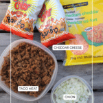 Top-down view of taco ingredients on a wooden surface: two bags of BBQ corn chips, a pack of shredded cheddar cheese, a container of cooked taco meat, and a small container of chopped onions.