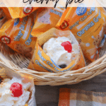 A basket filled with Bear Cub Grahams snack bags, one opened and filled with cherry pie filling, whipped cream, and a cherry on top. A plaid napkin and wooden table are visible beneath the basket.
