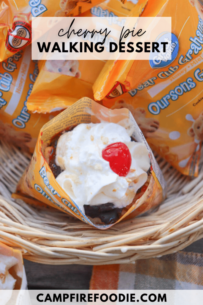 A waffle cone filled with cherry pie dessert, topped with whipped cream and a cherry, sits in a basket among Bear Club Grahams snack packs. Text overlay reads cherry pie WALKING DESSERT and CAMPFIREFOODIE.COM.