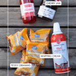 Five ingredients for a dessert, labeled: maraschino cherries, cherry jam, Teddy Grahams, whipping cream, and bear-shaped graham cracker packages, displayed on a wooden surface.