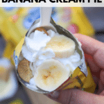 A hand holds an open mini cookie bag filled with sliced bananas, whipped cream, and cookies, with a plastic spoon inserted. The text reads walking dessert BANANA CREAM PIE and campfirefoodie.com.