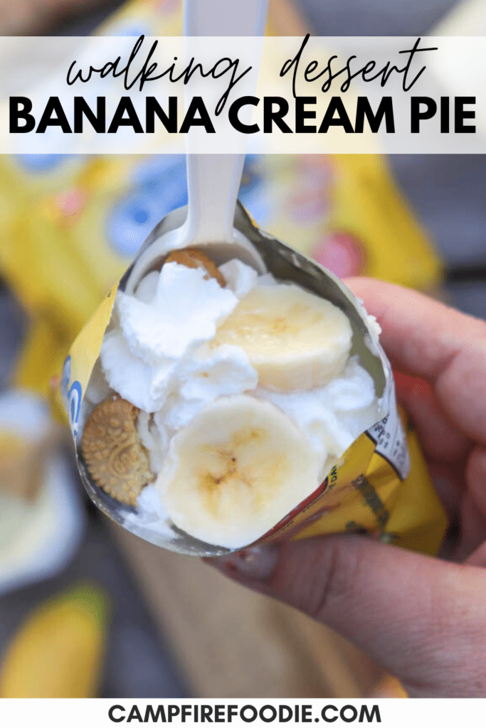 A hand holds an open mini cookie bag filled with sliced bananas, whipped cream, and cookies, with a plastic spoon inserted. The text reads walking dessert BANANA CREAM PIE and campfirefoodie.com.