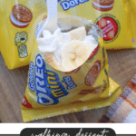 An opened Golden Oreo Mini bag is filled with whipped cream and banana slices, with a white plastic spoon inside. The bag sits on a wooden surface, next to more unopened Oreo Mini packs. Text reads: walking dessert BANANA CREAM PIE campfirefoodie.com.