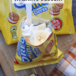 An open bag of Golden Oreo Mini cookies filled with whipped cream and banana slices sits on a cutting board with more cookies, a banana, and a plaid napkin nearby. Text reads “banana cream pie walking dessert.”.