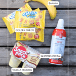 A banana, three packs of Golden Oreo Minis, a can of whipping cream, and two cups of vanilla pudding are arranged on a wooden surface with each item labeled.
