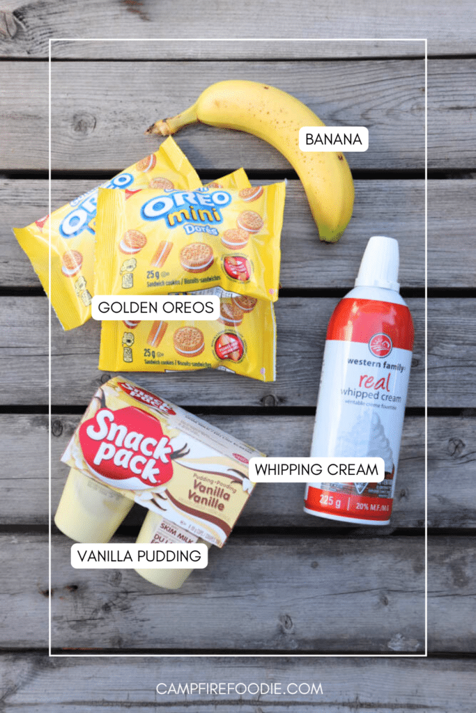 A banana, three packs of Golden Oreo Minis, a can of whipping cream, and two cups of vanilla pudding are arranged on a wooden surface with each item labeled.
