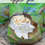 A bag of animal crackers is opened and filled with apple pie filling, topped with whipped cream and crushed crackers, sitting on a wicker surface. Text reads: Walking Dessert Apple Pie. Website: campfirefoodie.com.