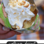 A hand holds a spoon digging into a snack bag filled with apple pie filling, topped with whipped cream and crushed cookies. Text reads: walking dessert APPLE PIE campfirefoodie.com.