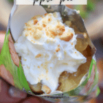 A close-up of a hand holding an open snack bag filled with apple pie filling, topped with whipped cream and crumbly bits. A plastic spoon is scooping into the dessert. Text reads, Walking Dessert Apple Pie and campfirefoodie.com.