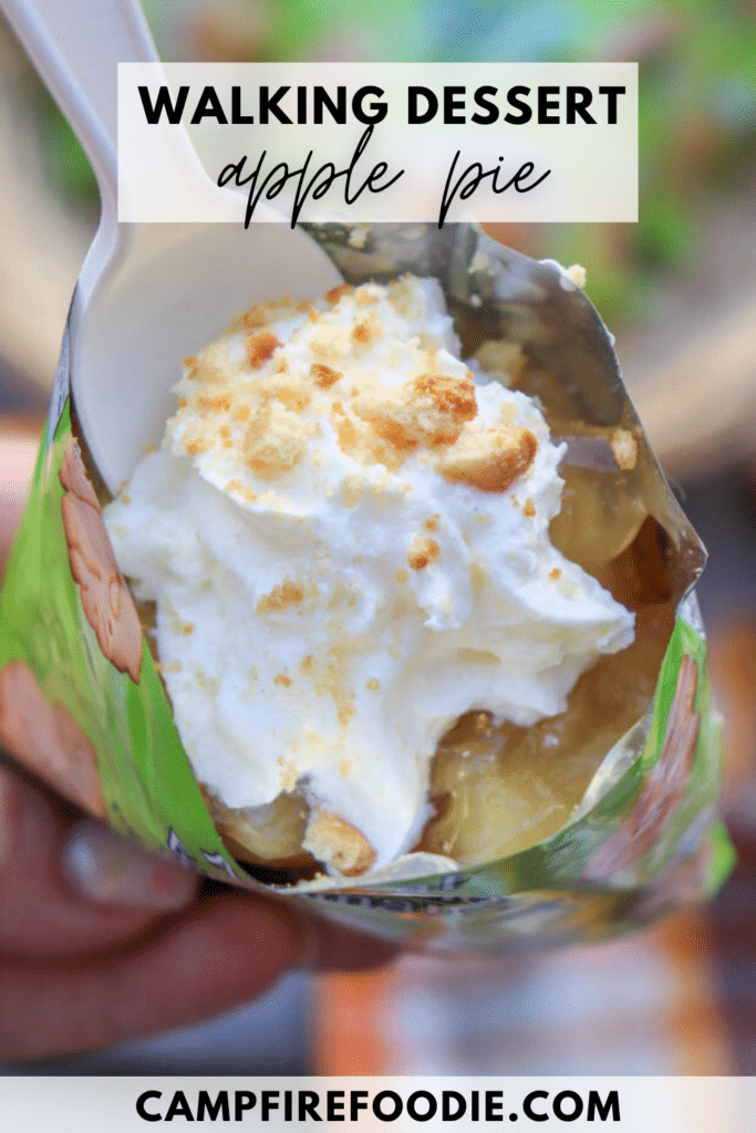 A close-up of a hand holding an open snack bag filled with apple pie filling, topped with whipped cream and crumbly bits. A plastic spoon is scooping into the dessert. Text reads, Walking Dessert Apple Pie and campfirefoodie.com.