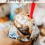 A hand holds an open snack bag filled with smores ingredients—marshmallow, chocolate, and graham cracker crumbs—topped with whipped cream and a red spoon. Text overlay reads: WALKING smores recipe.