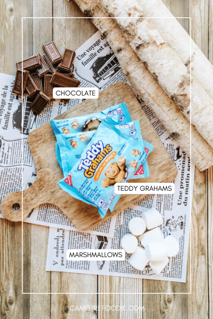 A wooden board holds Teddy Grahams, with chocolate pieces and marshmallows nearby. The items sit on newspaper atop a rustic wooden surface with a birch log. Labels identify chocolate, Teddy Grahams, and marshmallows.