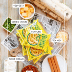 Top-down view of a wooden table with labeled ingredients: Velveeta cubes, green onions, Funyuns chips, shredded Tex Mex cheese, sour cream, chili, and hot dogs, along with birch logs and newspaper.